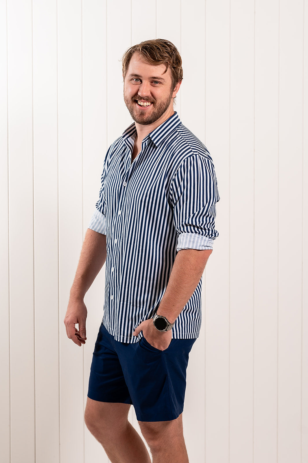 A smiling man wearing a blue and white vertically striped button-down shirt with rolled-up sleeves and navy blue shorts, standing against a white vertical-paneled wall.