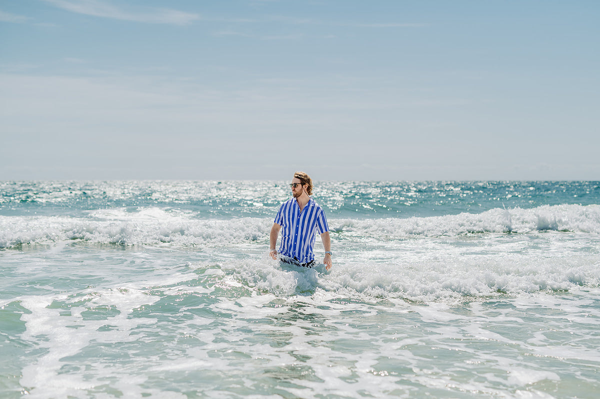 Man walking through the shallows at the beach wearing a blue striped Nashie